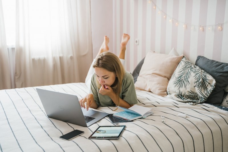 Una mujer está estirada en la cama, que utiliza como oficina en casa, trabajando con el ordenador portátil