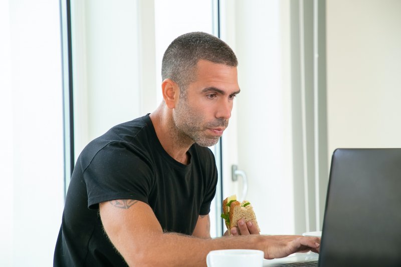 Un teletrabajador se pone a comer un bocadillo mientras utiliza el ordenador portátil