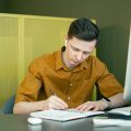 Un hombre que hace teletrabajo escribe en una libreta delante el ordenador