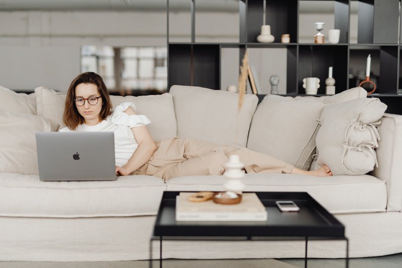 Una mujer que hace teletrabajo en su casa está estirada en el sofá con el ordenador portátil