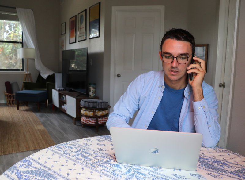 Un hombre hace trabajo flexible en el comedor de su casa, habla por el teléfono móvil mientras consulta el ordenador portátil